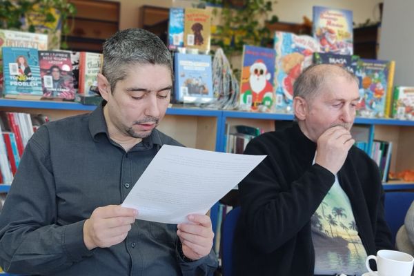 Mieszkańcy siedza i słuchaja tekstu czytanego.