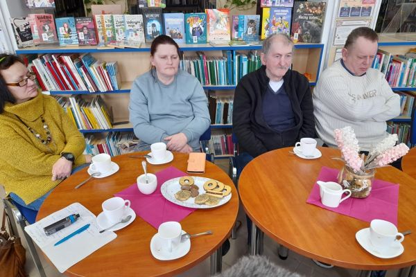 Mieszkańcy słuchają czytanego tekstu siedząc w bibliotece przy stolikach.