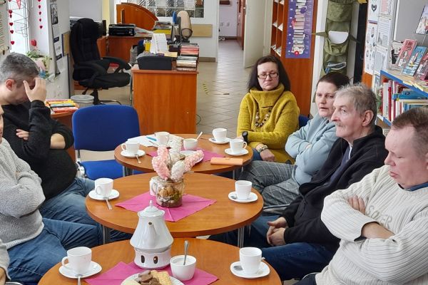 Mieszkańcy słuchają czytanego tekstu siedząc w bibliotece przy stolikach.