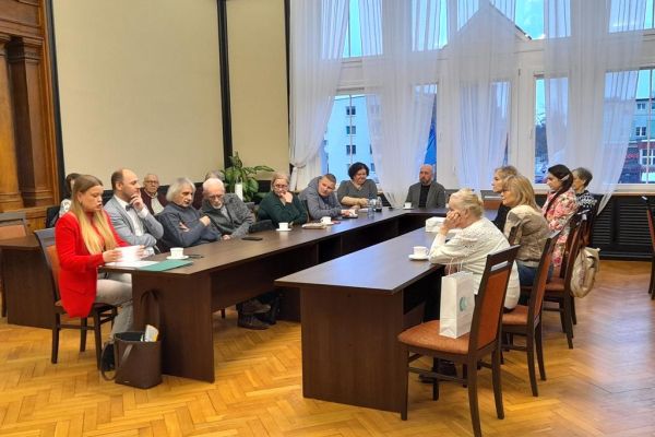 Mieszkanka siedzi przy długim stole wraz z innymi uczestnikami spotkania.