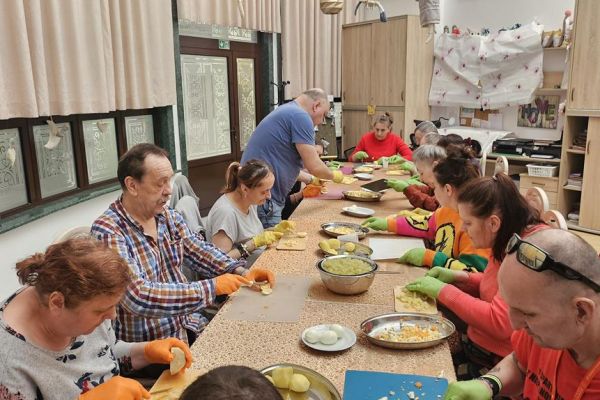 Mieszkańcy kroją produkty na słałatkę przy dużym stole.