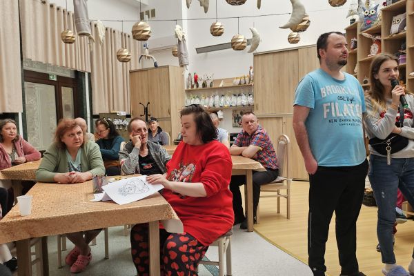 Jedni mieszkańcy siedzą przy stolikach i słuchają śpiewu a jedena osoba śpiewa na stojąco.