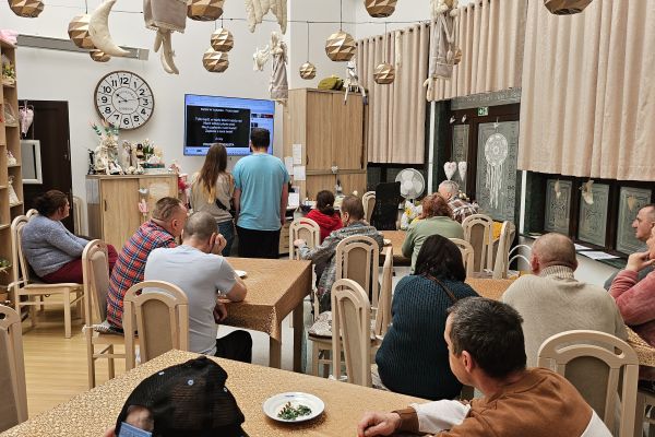 Jedni mieszkańcy siedzą przy stolikach i słuchają śpiewu a jedena osoba śpiewa na stojąco.