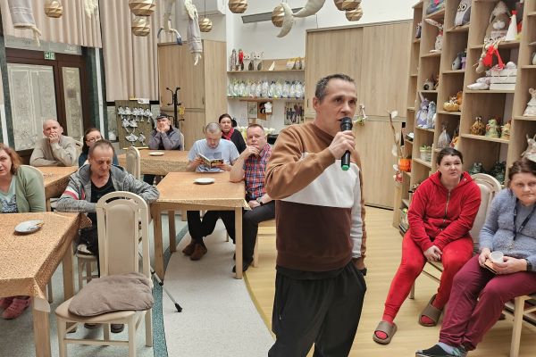 Jedni mieszkańcy siedzą przy stolikach i słuchają śpiewu a jedena osoba śpiewa na stojąco.