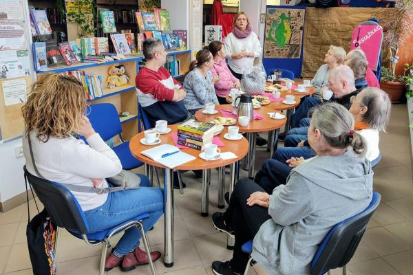 Mieszkańcy siedzą przy stolikach w bibliotece i słuchają czytanego tekstu.