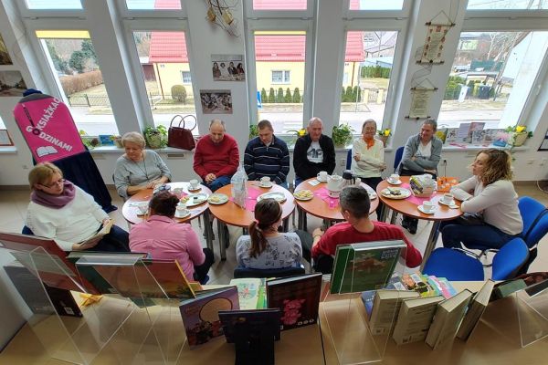 Mieszkańcy siedzą przy stolikach w bibliotece i słuchają czytanego tekstu.