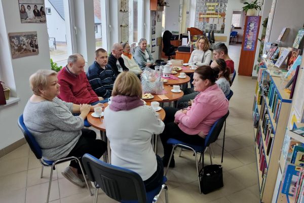 Mieszkańcy siedzą przy stolikach w bibliotece i słuchają czytanego tekstu.