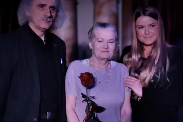 Mieszkanka trzymająca róże i dwoje uczestników spotkania stoi na scenie.