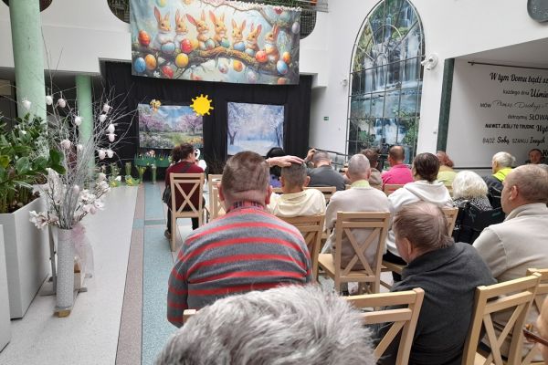 Mieszkańcy oglądfaja występ dzieci siedząc na krzesełkach w rzędach na holu.