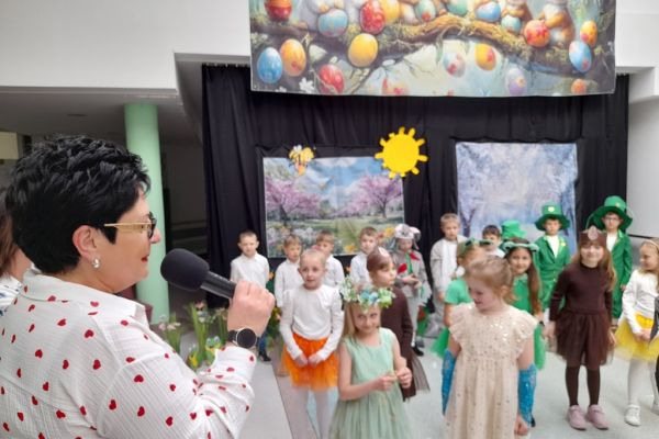 Dzieci i przed kotarą na holu Domu, a  Panią Dyrektor przemawia do mikrofonu.