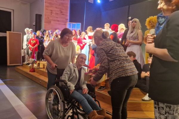 Mieszkanka siedząca na wózku inwalidzkim wręcza prezent Pani ze szkoły.