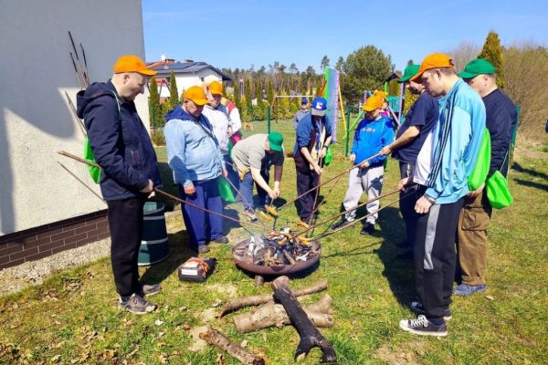 Mieszkańcy pieką kiełbaski w ognisku.