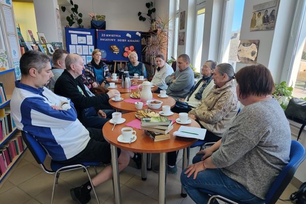 Mieszkańcy siedzą w bibliotece przy stolikach i słuchają tekstu czytanego.