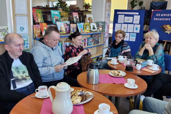 Mieszkańcy siedzą w bibliotece przy stolikach i słuchają tekstu czytanego.