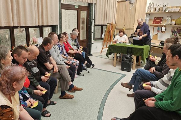 Mieszkańcy siedzą na krzesełkach w kole i odgadują piosenki.