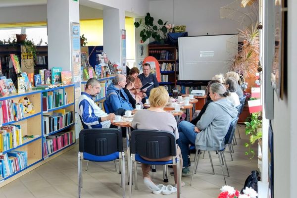 Mieszkańcy słuchaja tekstu czytanego siedząc przy stolikach w bibliotece.