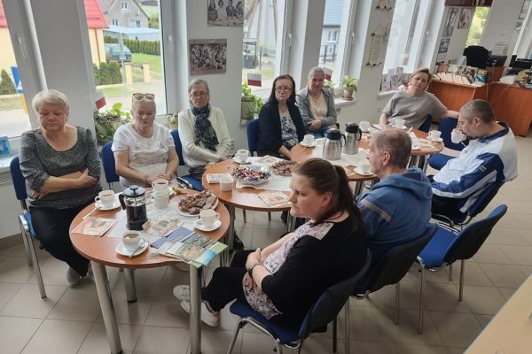 Mieszkańcy słuchaja tekstu czytanego siedząc przy stolikach w bibliotece.