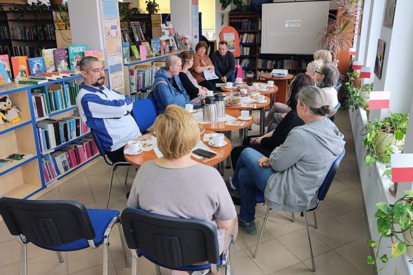 Mieszkańcy słuchaja tekstu czytanego siedząc przy stolikach w bibliotece.
