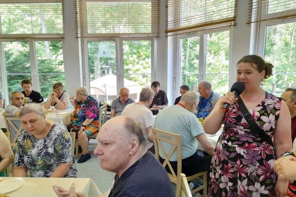 Mieszkańcy siedzą na stołówce przy stolikach i częstują się poczęstunkiem, jedna mieszkanka przemawia do mikrofonu stojąc.