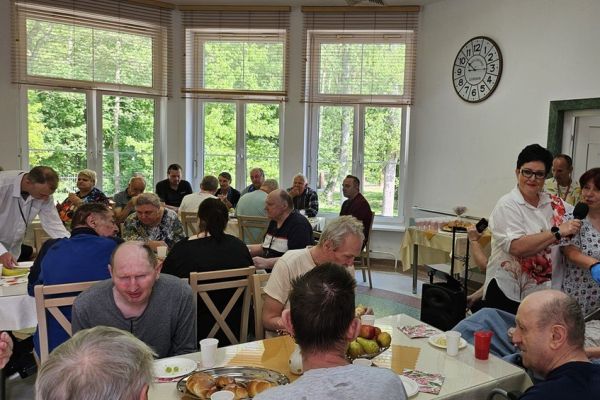 Mieszkańcy siedzą na stołówce przy stolikach i częstują się poczęstunkiem, jedna mieszkanka przemawia do mikrofonu stojąc.