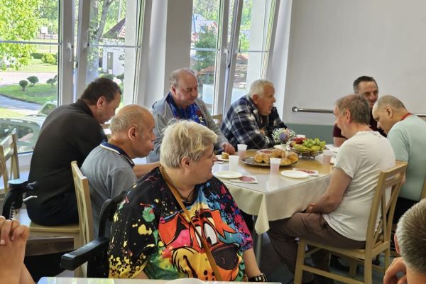 Mieszkańcy siedzą na stołówce przy stolikach i częstują się poczęstunkiem.