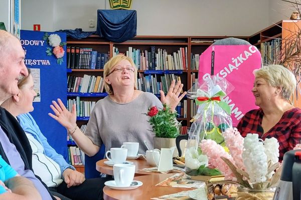 Mieszkańcy siedzą przy stolikach i słuchają czytanego tekstu przez jedną osobę.