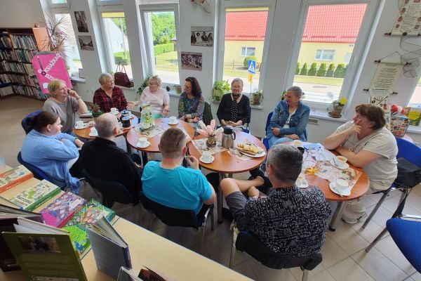 Mieszkańcy siedzą przy stolikach i słuchają czytanego tekstu przez jedną osobę.