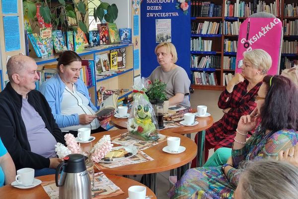 Mieszkańcy siedzą przy stolikach i słuchają czytanego tekstu przez jedną osobę.