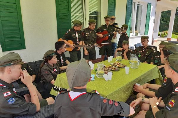 Harcerze graja na gitarach dla mieszkańców na tarasie domu.