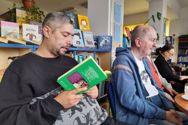 Mieszkaniec czyta książkę przy stoliku a inni uczestnicy słuchają.