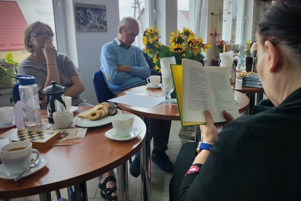 Mieszkanka czyta książkę przy stoliku a inni uczestnicy słuchają.