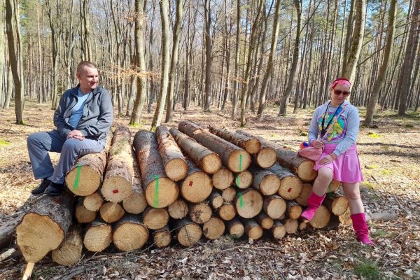 Dwoje Mieszkańców siedzi na kłodach ściętych drzew w lesie.