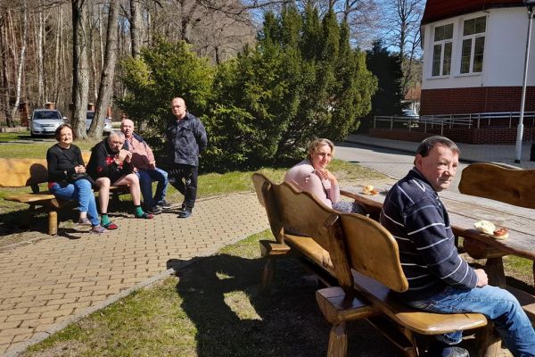 Mieszkańcy odpoczywają na ławkach przy domu pomocy i jedzą kiełbaskę z grilla.