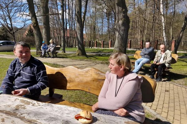 Mieszkańcy odpoczywają na ławkach przy domu pomocy i jedzą kiełbaskę z grilla.
