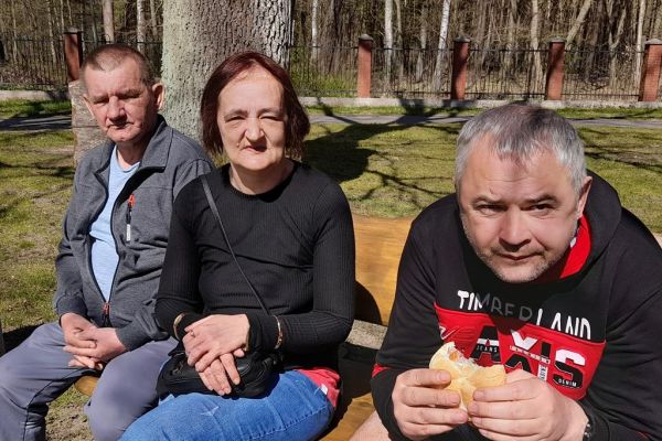 Mieszkańcy odpoczywają na ławkach przy domu pomocy i jedzą kiełbaskę z grilla.