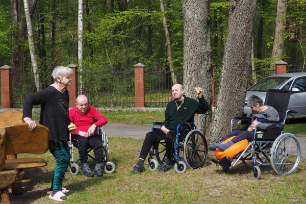 Mieszkańcy na wózkach spożywają kiełbaskę z grilla i odpoczywają na słoneczku.