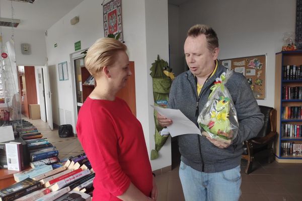 Mieszkaniec czyta życzenia z okazji Dnia bibliotekarza pani z biblioteki.
