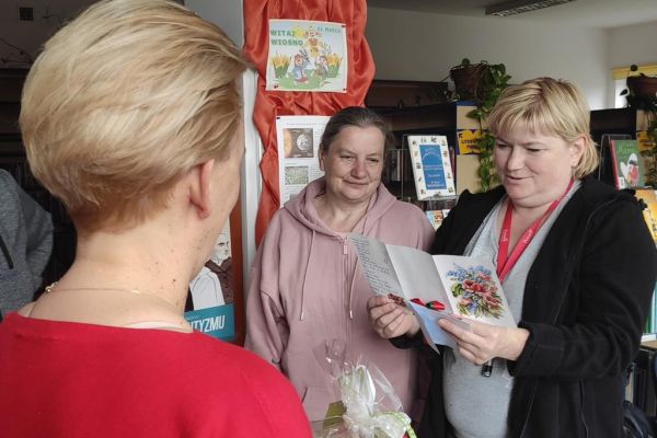 Terapetka zajęciowa czyta wiersz z okazji Dnia bibliotekarza dla pani z biblioteki.