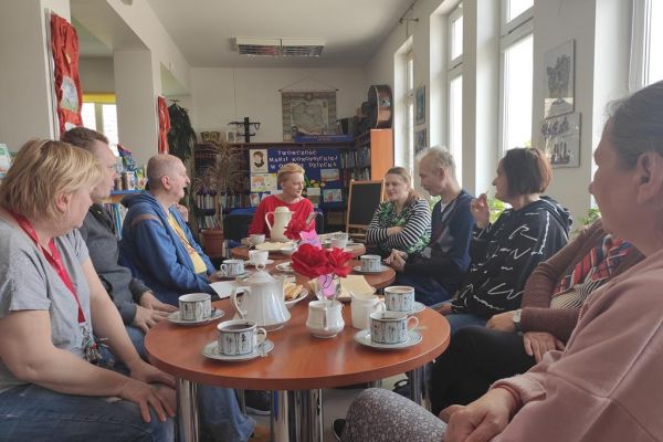 Mieszkańcy naprzemiennie opowiadają o przeczytanych książkach oraz słuchają wypowiedzi pani bibliotekarki siedząc przy stolikach.