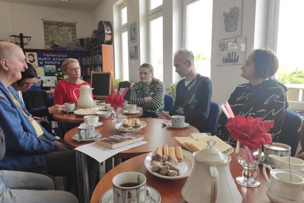 Mieszkańcy naprzemiennie opowiadają o przeczytanych książkach oraz słuchają wypowiedzi pani bibliotekarki siedząc przy stolikach.