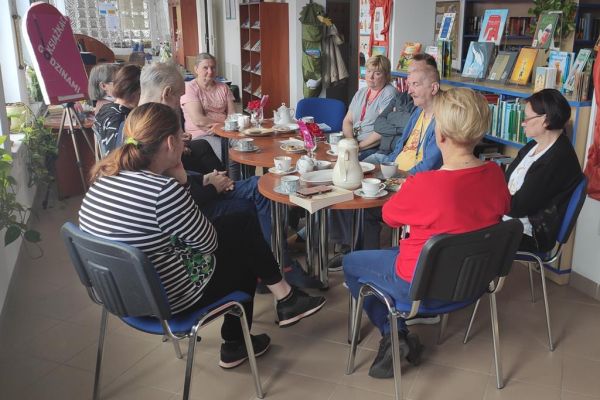 Mieszkańcy naprzemiennie opowiadają o przeczytanych książkach oraz słuchają wypowiedzi pani bibliotekarki siedząc przy stolikach.
