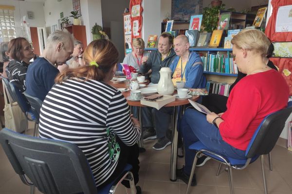Mieszkańcy naprzemiennie opowiadają o przeczytanych książkach oraz słuchają wypowiedzi pani bibliotekarki siedząc przy stolikach.
