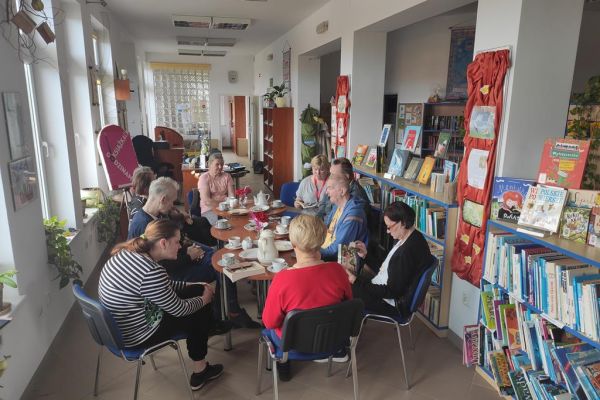 Mieszkańcy naprzemiennie opowiadają o przeczytanych książkach oraz słuchają wypowiedzi pani bibliotekarki siedząc przy stolikach.