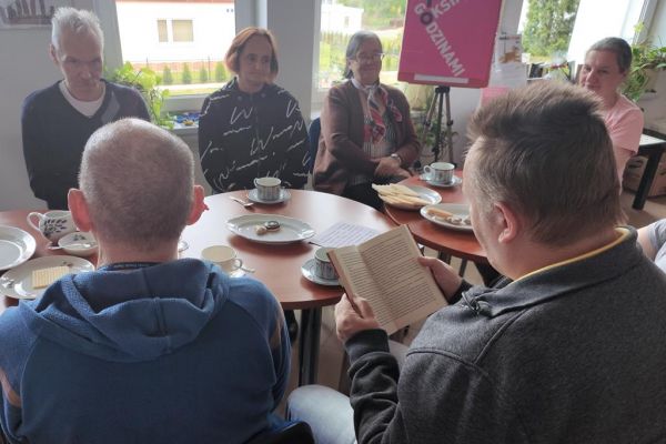 Mieszkańcy naprzemiennie opowiadają o przeczytanych książkach oraz słuchają wypowiedzi pani bibliotekarki siedząc przy stolikach.