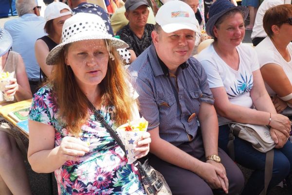 Mieszkańcy siedzą na ławeczce podczas wydarzenia słuchają zespołów folklorystycznych , a jedna z mieszkanek spożywa frytki.