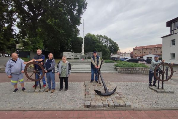 Mieszkańcy pozują do zdjęcia przy pomniku Ludziom morza trzymając kotwicę i koła sterowe.