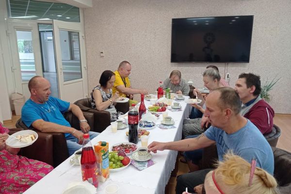 Mieszkańcy siędzą przy stołach wraz z jubilatem i kosztują poczęstunek.