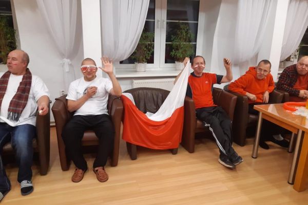 Mieszkańcy w strojach barwy biało - czerwonej oglądają mecz w kawiarence i kibicują.