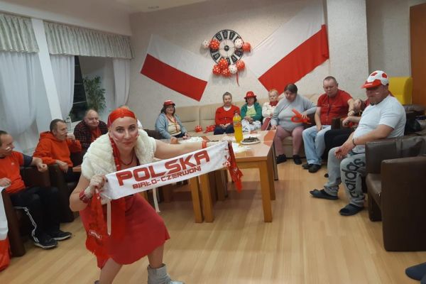 Mieszkańcy w strojach barwy biało - czerwonej oglądają mecz w kawiarence i kibicują.