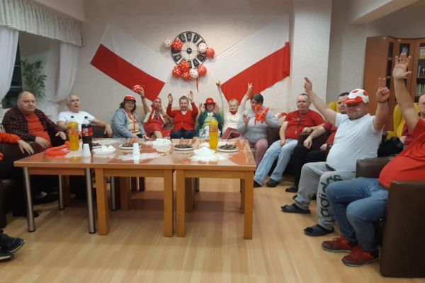 Mieszkańcy w strojach barwy biało - czerwonej oglądają mecz w kawiarence i kibicują.